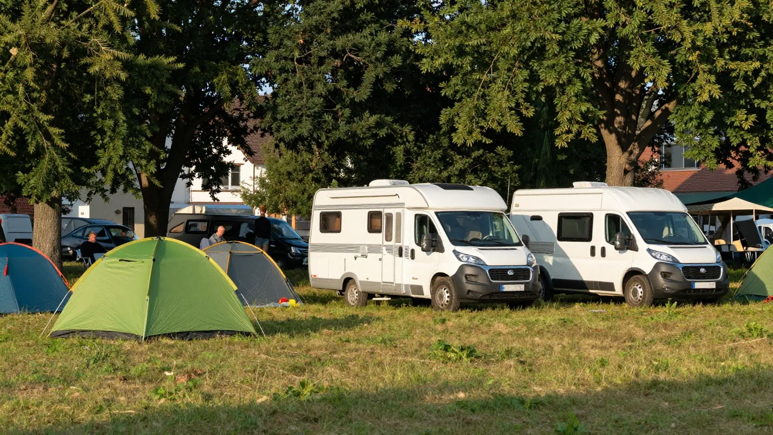 Eine Campingwiese am Nachmittag mit einem grünen Kuppelzelt, einem weißen Wohnwagen und einem kleinen Campingbus unter hohen Bäumen