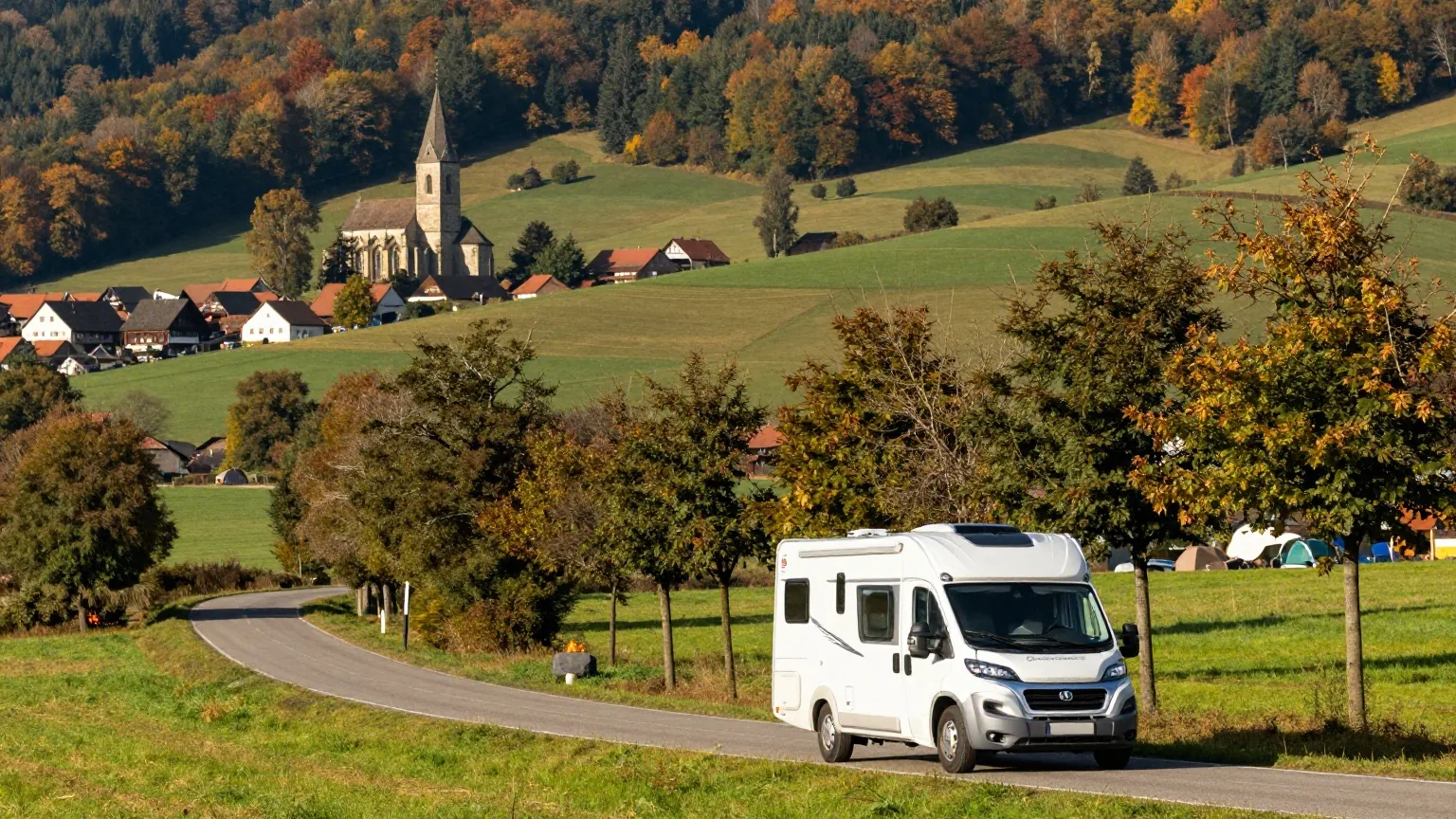 Ein weißes Wohnmobil auf einer ruhigen Allee durch hügelige Landschaft im Frühherbst, in der Ferne ein Dorf mit romanischer Kirche